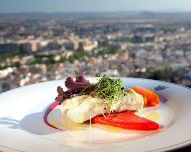 El plato de pescado gourmet, las verduras vibrantes y el paisaje urbano de fondo capturan perfectamente la esencia de Granada en el Hotel Alhambra Palace en el entorno "Almuerzo romántico a la carta". - ForTwo - Planes para parejas