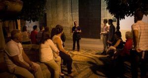 Un guía de "Tour guiado por los misterios de Córdoba" acompaña a un grupo sentado en un camino de piedra bajo el cielo nocturno, cerca de un gran muro. - ForTwo - Planes para parejas
