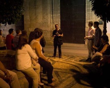Un guía de "Tour guiado por los misterios de Córdoba" acompaña a un grupo sentado en un camino de piedra bajo el cielo nocturno, cerca de un gran muro. - ForTwo - Planes para parejas
