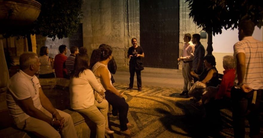 Un guía de "Tour guiado por los misterios de Córdoba" acompaña a un grupo sentado en un camino de piedra bajo el cielo nocturno, cerca de un gran muro. - ForTwo - Planes para parejas