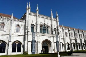 Un edificio histórico de piedra blanca con arcos y agujas ornamentados bajo un cielo azul despejado. Los carteles a la entrada guían a los visitantes para explorar estos monumentos, accesibles con la Lisboa Card: tarjeta con descuentos para acceder a los monumentos. - ForTwo - Planes para parejas