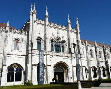 Un edificio histórico de piedra blanca con arcos y agujas ornamentados bajo un cielo azul despejado. Los carteles a la entrada guían a los visitantes para explorar estos monumentos, accesibles con la Lisboa Card: tarjeta con descuentos para acceder a los monumentos. - ForTwo - Planes para parejas