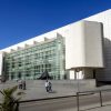 Un edificio moderno blanco con fachada de cristal, como la Entrada al Museo de Arte Contemporáneo (Barcelona), y gente caminando delante bajo un cielo azul claro. - ForTwo - Planes para parejas