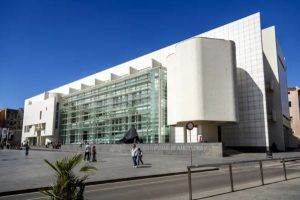 Un edificio moderno blanco con fachada de cristal, como la Entrada al Museo de Arte Contemporáneo (Barcelona), y gente caminando delante bajo un cielo azul claro. - ForTwo - Planes para parejas