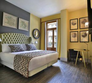 El NH Collection Colón en Valencia ofrece una elegante habitación con una cama grande adornada con cojines estampados, obras de arte enmarcadas, un espejo redondo y una ventana con encantadoras vistas a la calle. Disfrute de comodidad y sofisticación durante su estancia. - ForTwo - Planes para parejas