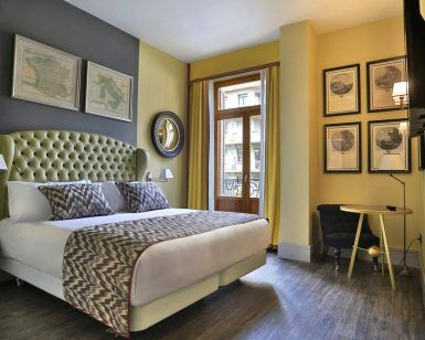 El NH Collection Colón en Valencia ofrece una elegante habitación con una cama grande adornada con cojines estampados, obras de arte enmarcadas, un espejo redondo y una ventana con encantadoras vistas a la calle. Disfrute de comodidad y sofisticación durante su estancia. - ForTwo - Planes para parejas