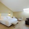 El elegante dormitorio minimalista del Hotel NH Collection Palacio de Oquendo en Cáceres incluye una cama grande, una iluminación suave en el tragaluz, un sofá oscuro y sillas alrededor de una mesa pequeña, todo ello realzado por ricos suelos de madera. - ForTwo - Planes para parejas