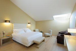 El elegante dormitorio minimalista del Hotel NH Collection Palacio de Oquendo en Cáceres incluye una cama grande, una iluminación suave en el tragaluz, un sofá oscuro y sillas alrededor de una mesa pequeña, todo ello realzado por ricos suelos de madera. - ForTwo - Planes para parejas