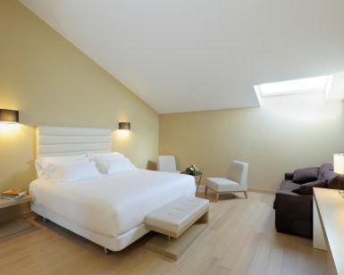 El elegante dormitorio minimalista del Hotel NH Collection Palacio de Oquendo en Cáceres incluye una cama grande, una iluminación suave en el tragaluz, un sofá oscuro y sillas alrededor de una mesa pequeña, todo ello realzado por ricos suelos de madera. - ForTwo - Planes para parejas