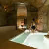Dos personas en traje de baño pasean por una piscina cubierta con paredes de piedra y techos abovedados, suavemente iluminada para crear un tranquilo ambiente de spa en el Pack noche + spa en el paraíso más exclusivo de agua termal milenaria (Barcelona). - ForTwo - Planes para parejas