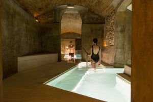 Dos personas en traje de baño pasean por una piscina cubierta con paredes de piedra y techos abovedados, suavemente iluminada para crear un tranquilo ambiente de spa en el Pack noche + spa en el paraíso más exclusivo de agua termal milenaria (Barcelona). - ForTwo - Planes para parejas