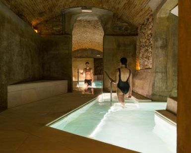 Dos personas en traje de baño pasean por una piscina cubierta con paredes de piedra y techos abovedados, suavemente iluminada para crear un tranquilo ambiente de spa en el Pack noche + spa en el paraíso más exclusivo de agua termal milenaria (Barcelona). - ForTwo - Planes para parejas