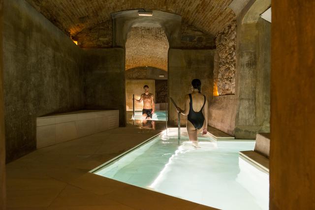 Dos personas en traje de baño pasean por una piscina cubierta con paredes de piedra y techos abovedados, suavemente iluminada para crear un tranquilo ambiente de spa en el Pack noche + spa en el paraíso más exclusivo de agua termal milenaria (Barcelona). - ForTwo - Planes para parejas