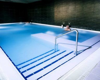 En Spa O2 Granada, la Oferta: Circuito ofrece una experiencia de relax en una piscina cubierta de agua cristalina y escaleras, rodeada de paredes de azulejos oscuros. - ForTwo - Planes para parejas