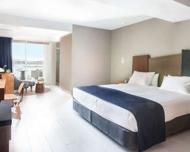 Experimente un lujo inigualable en una moderna habitación de hotel con una cama grande, escritorio de madera, silla, lámpara y amplios ventanales que dan a un balcón con serenas vistas a Cala Talamanca. Disfrute de esta exquisita escapada a Ibiza con la "Escapada de lujo en Cala Talamanca con desayuno". - ForTwo - Planes para parejas