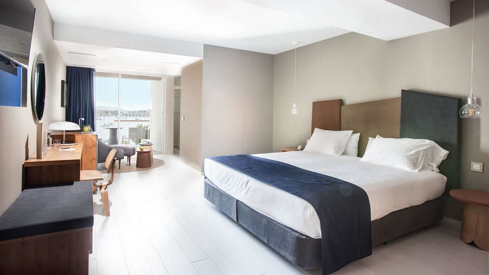 Experimente un lujo inigualable en una moderna habitación de hotel con una cama grande, escritorio de madera, silla, lámpara y amplios ventanales que dan a un balcón con serenas vistas a Cala Talamanca. Disfrute de esta exquisita escapada a Ibiza con la "Escapada de lujo en Cala Talamanca con desayuno". - ForTwo - Planes para parejas
