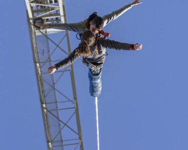 Una pareja atrevida se lanza al puenting desde una grúa contra el cielo azul claro de Barcelona, disfrutando del "Puenting en Barcelona en pareja". - ForTwo - Planes para parejas