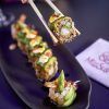 Los palillos sujetan un rollo de sushi de un plato negro con vibrantes piezas de sushi en Miss Sushi Granada; la servilleta descansa sobre la mesa oscura. - ForTwo - Planes para parejas