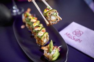 Los palillos sujetan un rollo de sushi de un plato negro con vibrantes piezas de sushi en Miss Sushi Granada; la servilleta descansa sobre la mesa oscura. - ForTwo - Planes para parejas