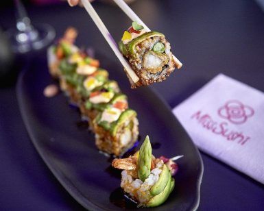 Los palillos sujetan un rollo de sushi de un plato negro con vibrantes piezas de sushi en Miss Sushi Granada; la servilleta descansa sobre la mesa oscura. - ForTwo - Planes para parejas