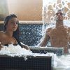 En el tranquilo Kumaras Spa en Umbrete, dos personas se relajan en una piscina de spa con cascada de agua; uno sentado mientras el otro permanece de pie bajo el refrescante flujo, saboreando la experiencia del Circuito Termal 90 minutos. - ForTwo - Planes para parejas