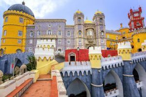 Ubicada en las vibrantes afueras de Lisboa, la "Excursión por los alrededores más turísticos de Lisboa" ofrece una visita a un colorido palacio con torres amarillas, rojas y grises adornadas con arcos y detalles intrincados bajo un cielo azul sereno, ideal para una excursión. - ForTwo - Planes para parejas