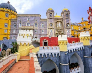Ubicada en las vibrantes afueras de Lisboa, la "Excursión por los alrededores más turísticos de Lisboa" ofrece una visita a un colorido palacio con torres amarillas, rojas y grises adornadas con arcos y detalles intrincados bajo un cielo azul sereno, ideal para una excursión. - ForTwo - Planes para parejas