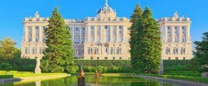 Explora el Palacio Real de Madrid, un palacio impresionante con una fachada detallada, rodeado de un jardín cuidadosamente mantenido, piscina reflectante y cielo azul claro. - ForTwo - Planes para parejas