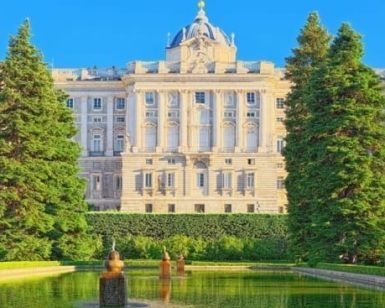 Explora el Palacio Real de Madrid, un palacio impresionante con una fachada detallada, rodeado de un jardín cuidadosamente mantenido, piscina reflectante y cielo azul claro. - ForTwo - Planes para parejas