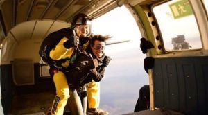 Dos personas sonríen mientras están bien sujetas a su paracaídas, disfrutando de un Salto tándem de paracaidismo con Skydive Empuriabrava mientras saltan en paracaídas desde un avión. - ForTwo - Planes para parejas