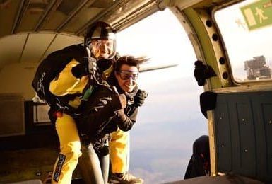 Dos personas sonríen mientras están bien sujetas a su paracaídas, disfrutando de un Salto tándem de paracaidismo con Skydive Empuriabrava mientras saltan en paracaídas desde un avión. - ForTwo - Planes para parejas