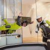 Dos personas disfrutan de la Experiencia en el Túnel de Viento en Madrid: una volando con un traje verde, la otra levantando el pulgar, ambas con casco. Una emocionante experiencia que desafía la gravedad. - ForTwo - Planes para parejas