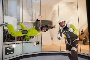 Dos personas disfrutan de la Experiencia en el Túnel de Viento en Madrid: una volando con un traje verde, la otra levantando el pulgar, ambas con casco. Una emocionante experiencia que desafía la gravedad. - ForTwo - Planes para parejas