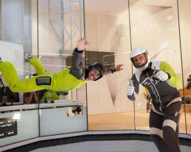 Dos personas disfrutan de la Experiencia en el Túnel de Viento en Madrid: una volando con un traje verde, la otra levantando el pulgar, ambas con casco. Una emocionante experiencia que desafía la gravedad. - ForTwo - Planes para parejas