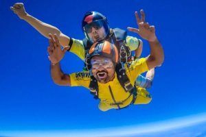 Dos personas saltando en paracaídas en tándem contra un cielo azul brillante, sonriendo ampliamente con equipos coloridos, personifican la emocionante aventura de "Salto en paracaídas con SkyDive Sevilla", una experiencia verdaderamente inolvidable. - ForTwo - Planes para parejas