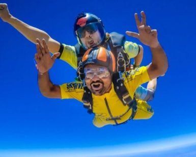 Dos personas saltando en paracaídas en tándem contra un cielo azul brillante, sonriendo ampliamente con equipos coloridos, personifican la emocionante aventura de "Salto en paracaídas con SkyDive Sevilla", una experiencia verdaderamente inolvidable. - ForTwo - Planes para parejas