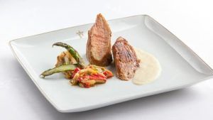 Un plato gourmet con dos suculentas carnes en salsa cremosa y verduras mixtas sobre un plato cuadrado blanco evoca las refinadas experiencias culinarias del Restaurante Torreorgaz Parador de Cáceres. - ForTwo - Planes para parejas