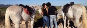 Una pareja comparte un beso entre dos caballos blancos ensillados en una ladera pintoresca durante el "Paseo a caballo por la sierra de Guadarrama con picnic para dos". - ForTwo - Planes para parejas