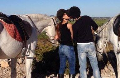 Una pareja comparte un beso entre dos caballos blancos ensillados en una ladera pintoresca durante el "Paseo a caballo por la sierra de Guadarrama con picnic para dos". - ForTwo - Planes para parejas
