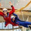 Dos personas experimentan la emoción del "Paracaidismo indoor en túnel de viento Madrid Fly", maniobrando con gracia en un túnel de viento mientras visten llamativos trajes, cascos y gafas de color rojo y azul. - ForTwo - Planes para parejas