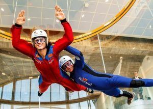 Dos personas experimentan la emoción del "Paracaidismo indoor en túnel de viento Madrid Fly", maniobrando con gracia en un túnel de viento mientras visten llamativos trajes, cascos y gafas de color rojo y azul. - ForTwo - Planes para parejas