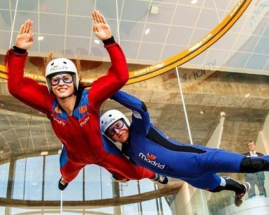 Dos personas experimentan la emoción del "Paracaidismo indoor en túnel de viento Madrid Fly", maniobrando con gracia en un túnel de viento mientras visten llamativos trajes, cascos y gafas de color rojo y azul. - ForTwo - Planes para parejas