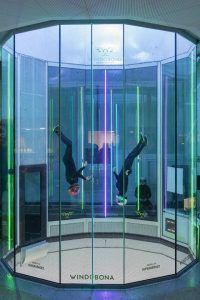 Dos personas con cascos saltan en paracaídas en el interior de Windobona Indoor Skydiving Madrid, en un túnel de viento cilíndrico con vibrantes luces verticales. - ForTwo - Planes para parejas