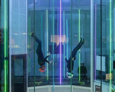 Dos personas con cascos saltan en paracaídas en el interior de Windobona Indoor Skydiving Madrid, en un túnel de viento cilíndrico con vibrantes luces verticales. - ForTwo - Planes para parejas