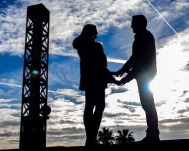 Una pareja de siluetas cogidas de la mano bajo un cielo nublado de Barcelona con árboles y estructuras al fondo ejemplifica maravillosamente la Sesión de fotografía romántica al aire libre (Barcelona). - ForTwo - Planes para parejas