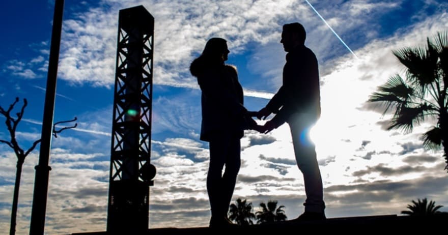 Una pareja de siluetas cogidas de la mano bajo un cielo nublado de Barcelona con árboles y estructuras al fondo ejemplifica maravillosamente la Sesión de fotografía romántica al aire libre (Barcelona). - ForTwo - Planes para parejas