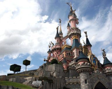 Un majestuoso castillo rosa con torres se yergue orgulloso bajo un cielo azul salpicado de nubes esponjosas, que recuerda una escena sacada directamente de una excursión a Disneyland París desde París. - ForTwo - Planes para parejas