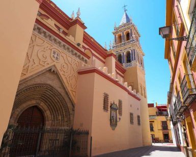 Una calle estrecha con una gran iglesia ornamentada y un campanario imponente bajo un cielo azul brillante, que ofrece el ambiente encantador del Barrio de Triana de Sevilla, como se experimenta en el "Tour gratuito por el barrio de Triana (Sevilla). - ForTwo - Planes para parejas