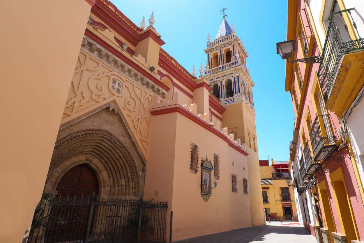 Una calle estrecha con una gran iglesia ornamentada y un campanario imponente bajo un cielo azul brillante, que ofrece el ambiente encantador del Barrio de Triana de Sevilla, como se experimenta en el "Tour gratuito por el barrio de Triana (Sevilla). - ForTwo - Planes para parejas