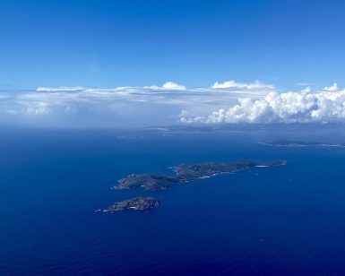Experimente la impresionante vista de una exuberante isla verde rodeada por el océano azul profundo bajo un cielo despejado con nubes dispersas, como si volara en una "Sobrevuela en avión las Islas Cíes (Vigo)", capturando el encanto encantador de las Islas Cíes cerca de Vigo. - ForTwo - Planes para parejas